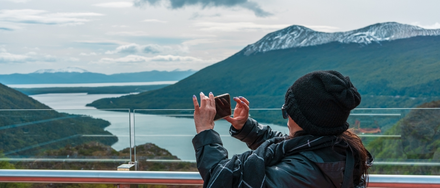 Persona con ropa de abrigo tomando una foto desde el mirador Paso Garibaldi, con vista panorámica a un lago rodeado de montañas boscosas y picos nevados bajo un cielo nublado.