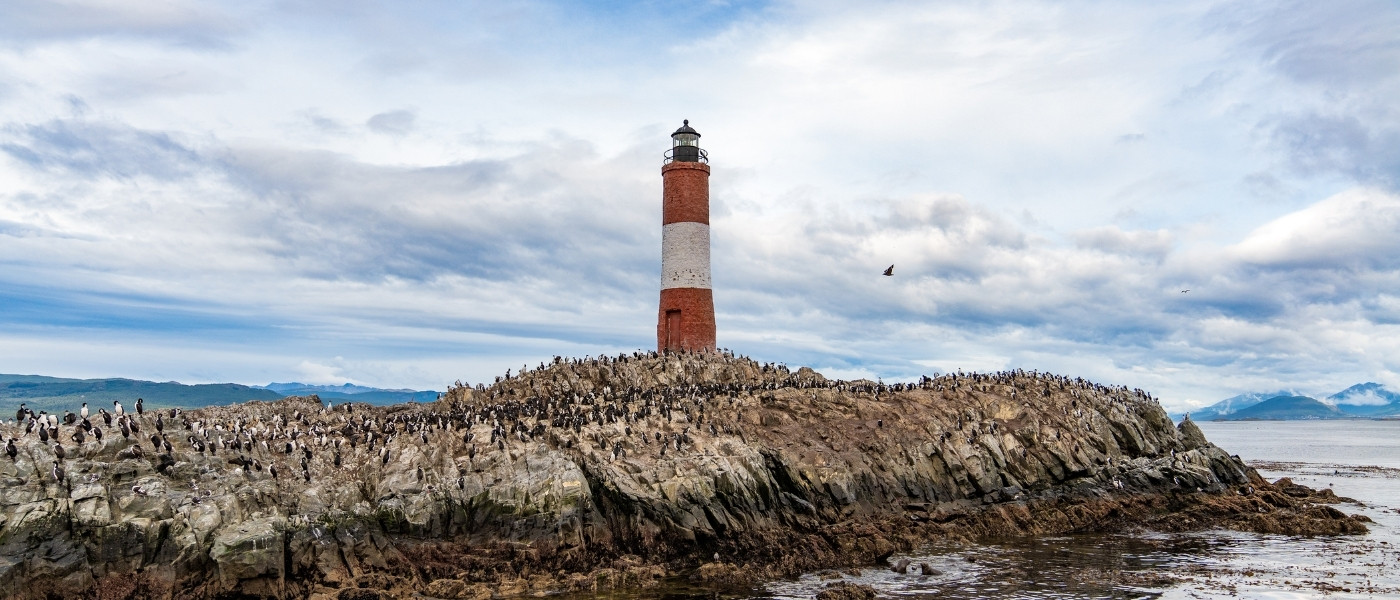 Isla rocosa poblada por lobos marinos y aves, con el faro Les Eclaireurs rojo y blanco en el centro, rodeada de aguas tranquilas y montañas al fondo bajo un cielo parcialmente nublado.