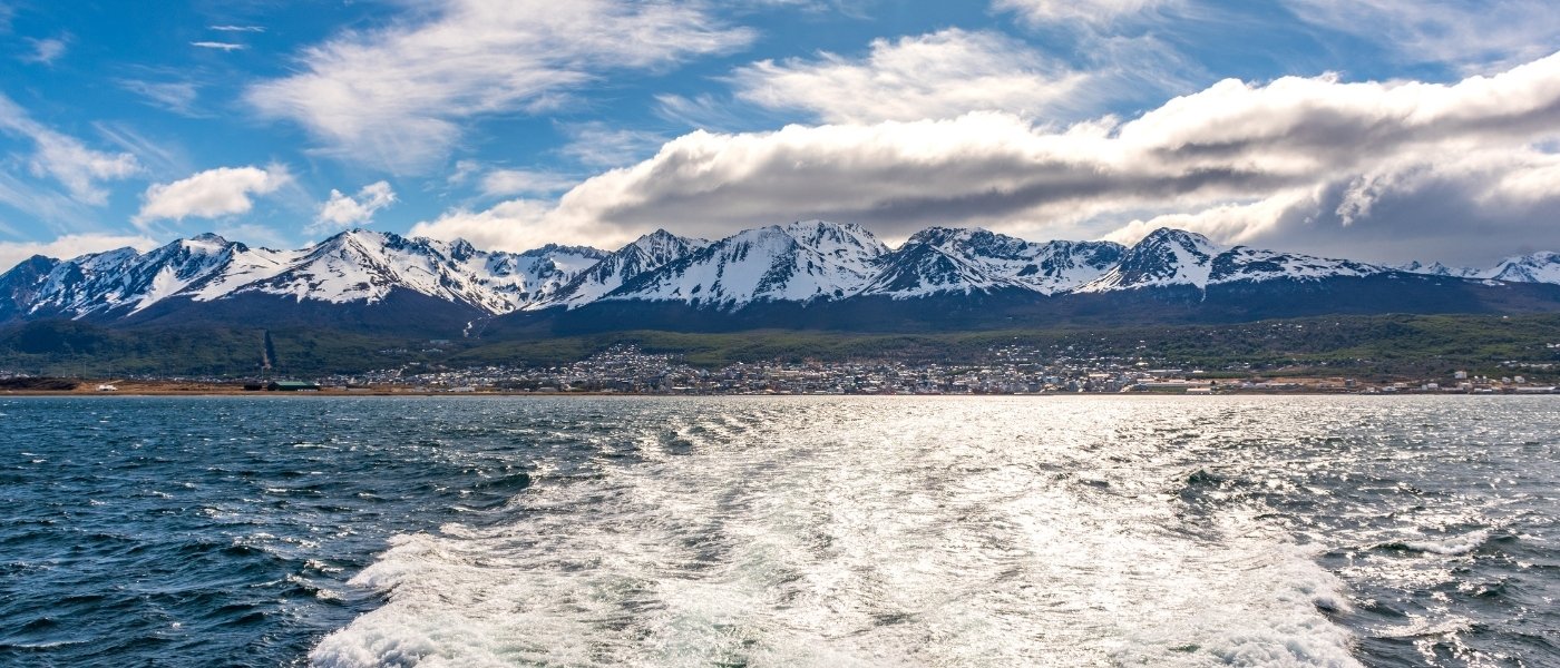 Vista desde una embarcación navegando por el Canal Beagle, con el mar en primer plano y la ciudad al pie de montañas nevadas bajo un cielo parcialmente nublado.