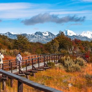 Dos excursionistas caminando por una pasarela de madera en el Parque Nacional Tierra del Fuego, rodeados de colores otoñales y montañas nevadas.