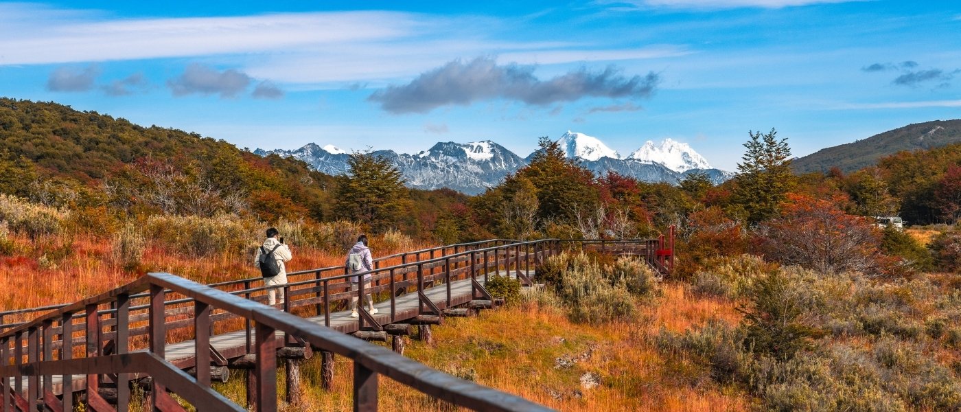 Dos excursionistas caminando por una pasarela de madera en el Parque Nacional Tierra del Fuego, rodeados de colores otoñales y montañas nevadas.