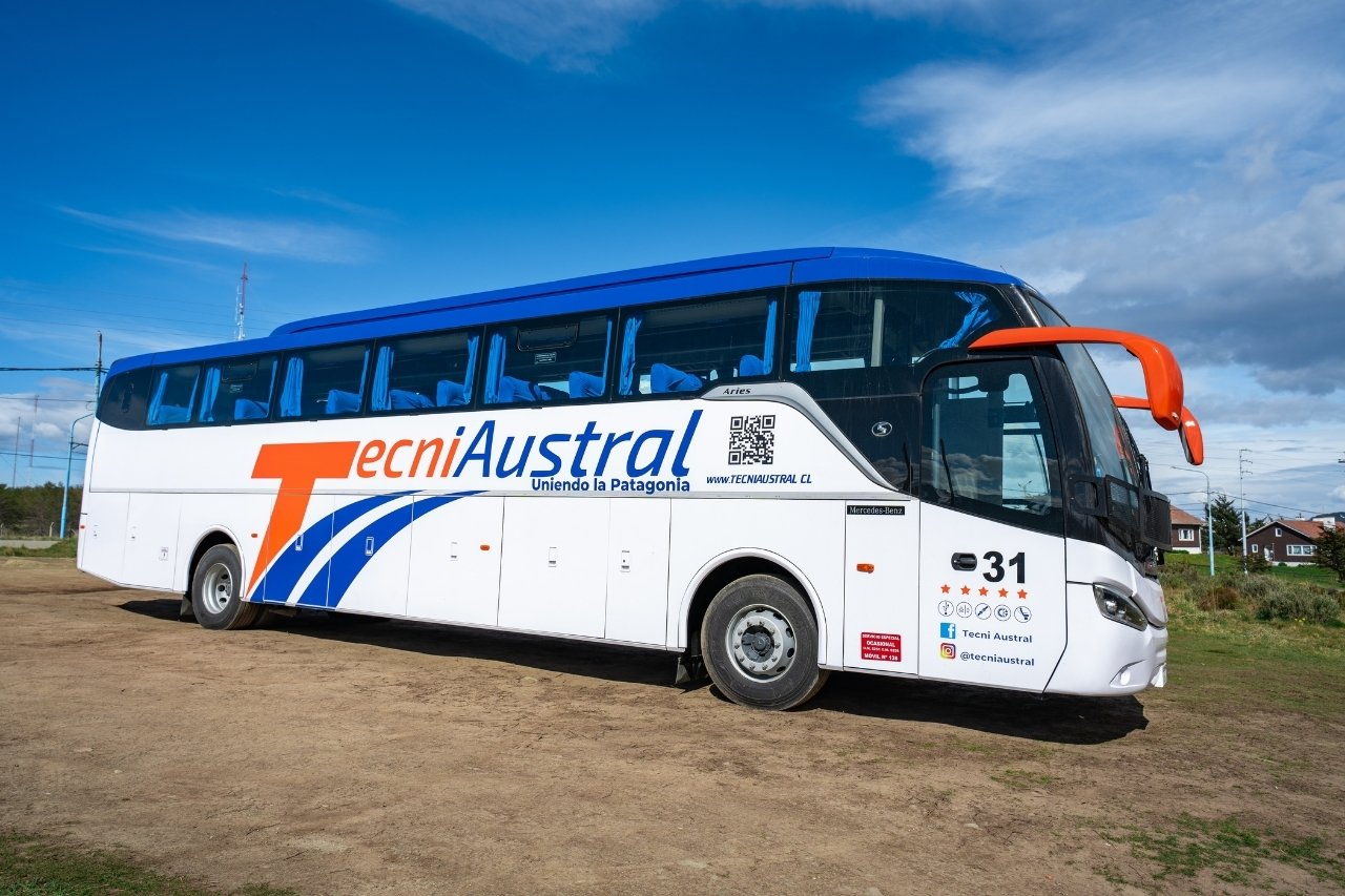 Autobús de larga distancia de la empresa TecniAustral, modelo Mercedes-Benz, estacionado en Ushuaia. Vista lateral derecha mostrando la marca y colores blanco, naranja y azul.