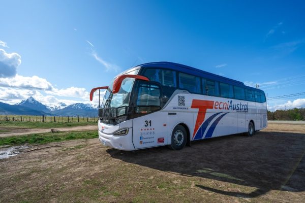 Frente diagonal del autobús TecniAustral en Ushuaia, destacando los espejos naranja, el logo Mercedes-Benz y la cordillera nevada de fondo.