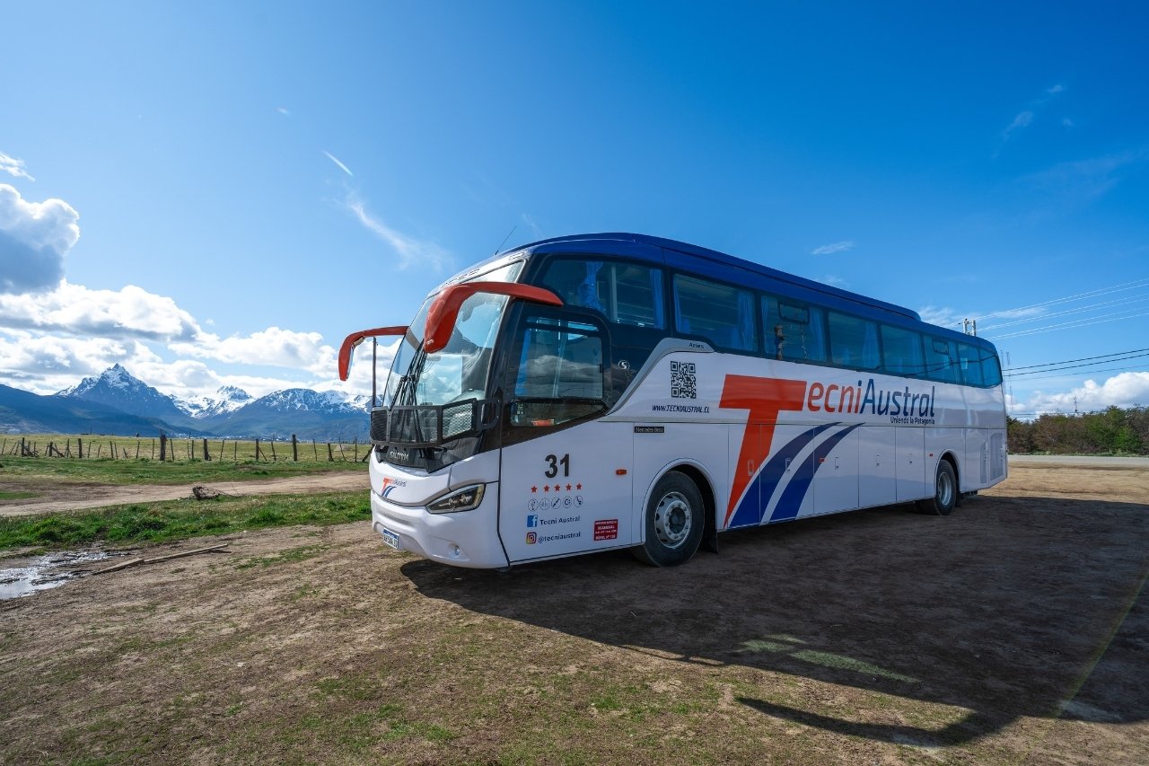 Frente diagonal del autobús TecniAustral en Ushuaia, destacando los espejos naranja, el logo Mercedes-Benz y la cordillera nevada de fondo.