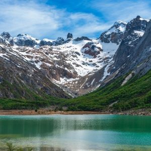 La Laguna Esmeralda en Ushuaia, rodeada de bosques verdes y las majestuosas montañas andinas con picos nevados bajo un cielo azul con nubes.