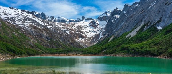 La Laguna Esmeralda en Ushuaia, rodeada de bosques verdes y las majestuosas montañas andinas con picos nevados bajo un cielo azul con nubes.
