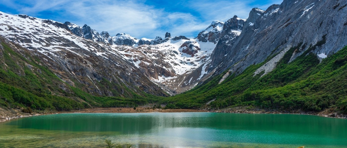 Paisaje de la Laguna Esmeralda con aguas turquesa rodeadas de montañas escarpadas parcialmente nevadas y bosques verdes bajo un cielo despejado con algunas nubes.