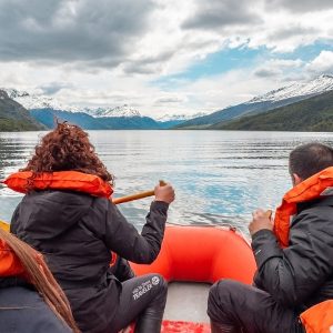 Turistas navegando en un bote inflable naranja por el Lago Roca (Lago Acigami) en el Parque Nacional Tierra del Fuego, Ushuaia. Se ven montañas nevadas y bosques nativos bajo un cielo nublado.