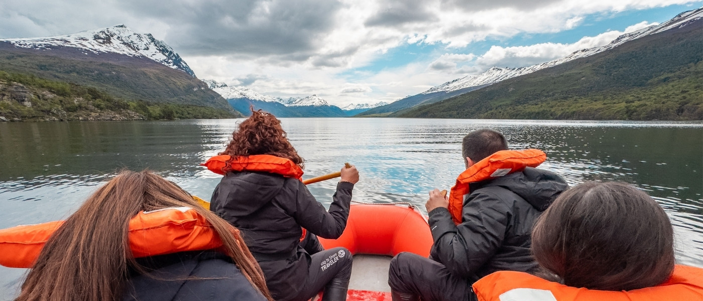 Turistas navegando en un bote inflable naranja por el Lago Roca (Lago Acigami) en el Parque Nacional Tierra del Fuego, Ushuaia. Se ven montañas nevadas y bosques nativos bajo un cielo nublado.