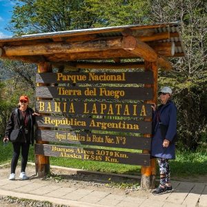 Dos mujeres posando sonrientes junto al icónico cartel de madera de la Bahía Lapataia en el Parque Nacional Tierra del Fuego, Ushuaia.