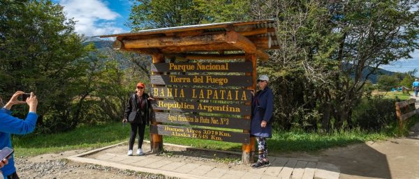 Dos mujeres posando sonrientes junto al icónico cartel de madera de la Bahía Lapataia en el Parque Nacional Tierra del Fuego, Ushuaia.