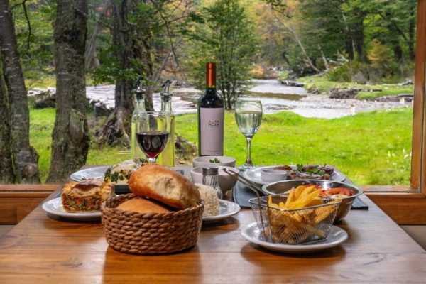 Almuerzo servido en una mesa de madera frente a un ventanal con vistas al bosque fueguino y el río Tristen en Ushuaia.