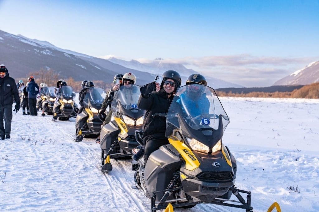 Un grupo de personas en una excursión guiada en motos de nieve por el Park Austral se toma una selfie sonriendo, con montañas nevadas al fondo.