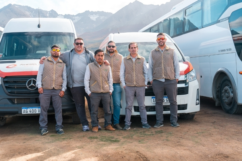 El equipo de choferes de Aswalek Expediciones posando junto a la flota de combis Mercedes-Benz y Toyota en Ushuaia, con las montañas de los Andes de fondo.