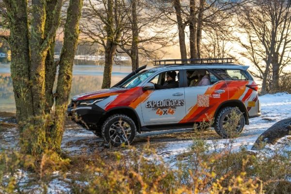 Una camioneta Toyota SW4 blanca con el logo de Aswalek Expediciones 4x4, equipada con portaequipajes, estacionada en la orilla nevada del Lago Escondido en Ushuaia, rodeada de un bosque helado bajo el sol del amanecer.