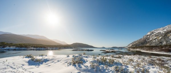 Paisaje soleado de Bahía Lapataia en invierno, con el mar en calma, montañas nevadas y la costa cubierta de nieve bajo un cielo azul despejado.