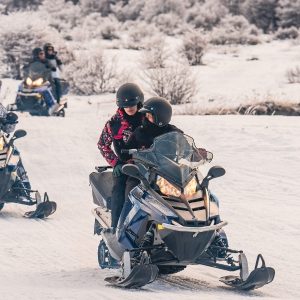 Un grupo de turistas con cascos y ropa de nieve conduce motos de nieve azules y blancas en fila india por un camino cubierto de nieve profunda en un valle andino de Ushuaia, bajo un cielo nublado parcial con bosques de lengas helados.