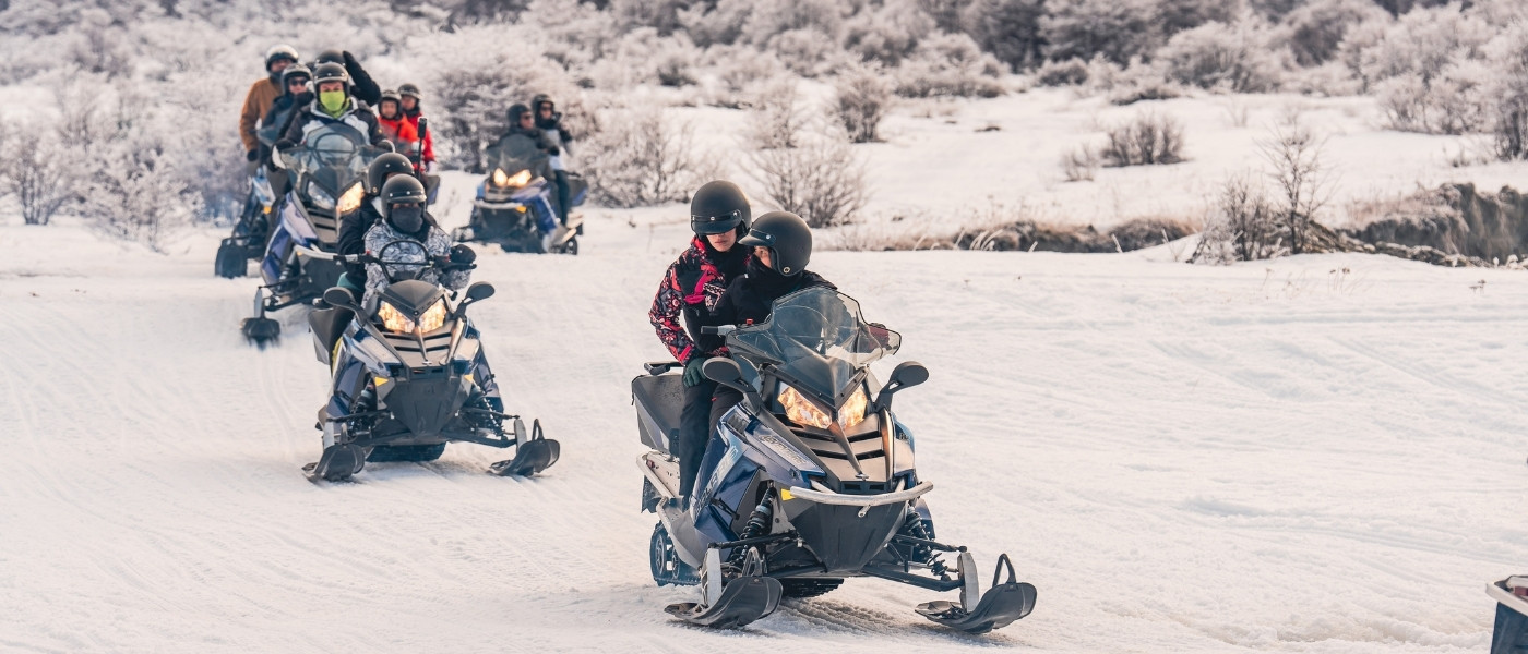 Un grupo de turistas con cascos y ropa de nieve conduce motos de nieve azules y blancas en fila india por un camino cubierto de nieve profunda en un valle andino de Ushuaia, bajo un cielo nublado parcial con bosques de lengas helados.