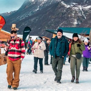 Grupo de turistas caminando por un sendero de nieve en un centro invernal de Ushuaia, con un hombre en primer plano mirando su smartphone, una pareja sonriendo detrás de él y montañas nevadas borrosas y cabañas de madera al fondo bajo un cielo nublado.