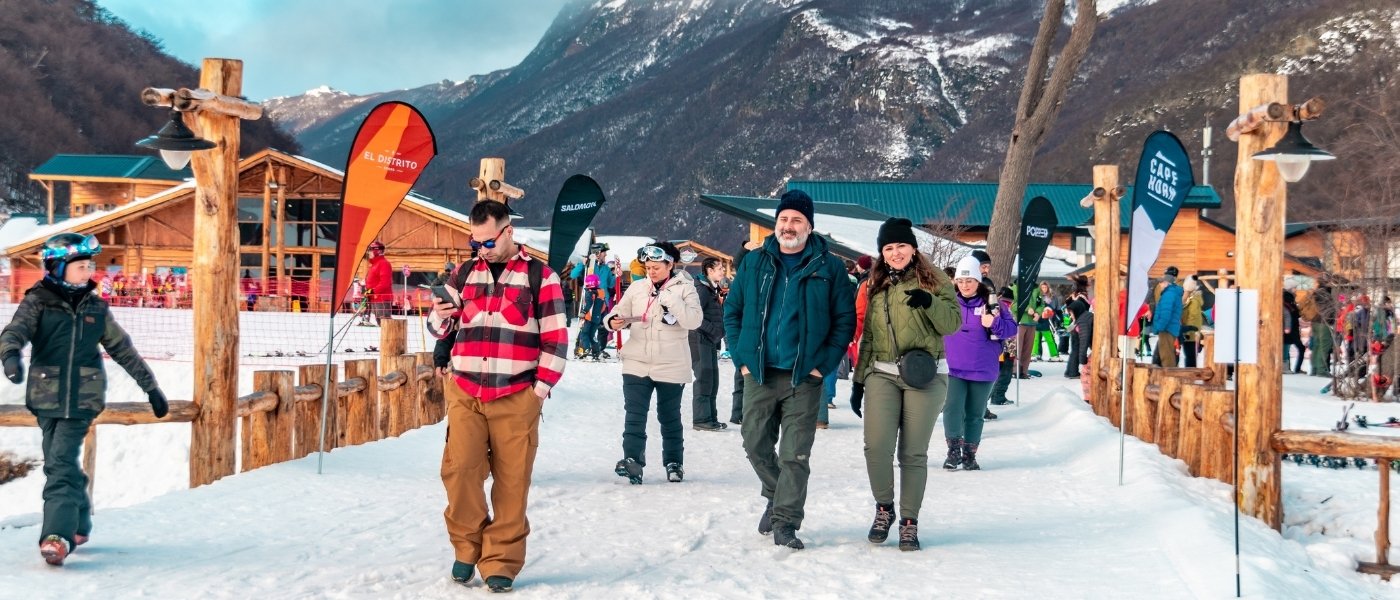 Grupo de turistas caminando por un sendero de nieve en un centro invernal de Ushuaia, con un hombre en primer plano mirando su smartphone, una pareja sonriendo detrás de él y montañas nevadas borrosas y cabañas de madera al fondo bajo un cielo nublado.