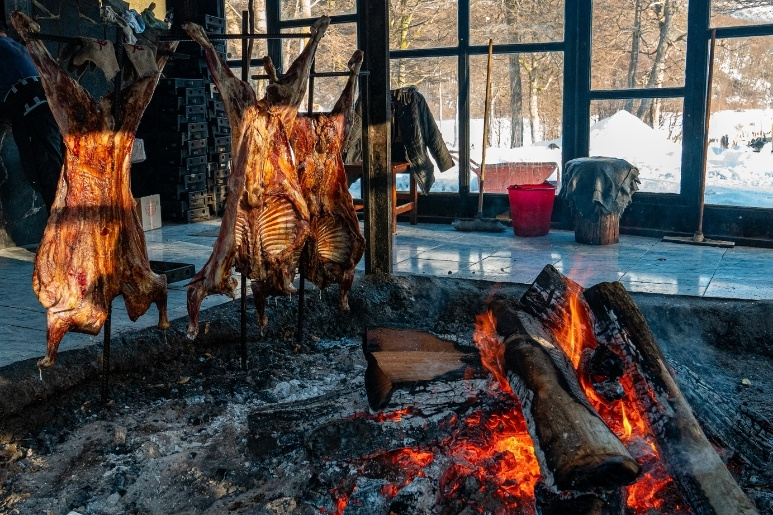 Tres corderos fueguinos enteros cocinándose lentamente al asador en cruz sobre un gran fogón de leña ardiendo dentro del restaurante rústico del centro invernal Las Cotorras en Ushuaia, con nieve visible a través de los ventanales al fondo.