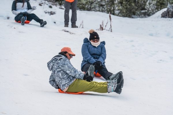 Dos turistas vestidas con ropa de invierno técnica, cascos y anteojos de sol sonríen mientras se deslizan rápido en un trineo naranja por una ladera de nieve profunda en el centro invernal Las Cotorras en Ushuaia. Otro turista en trineo está más arriba.