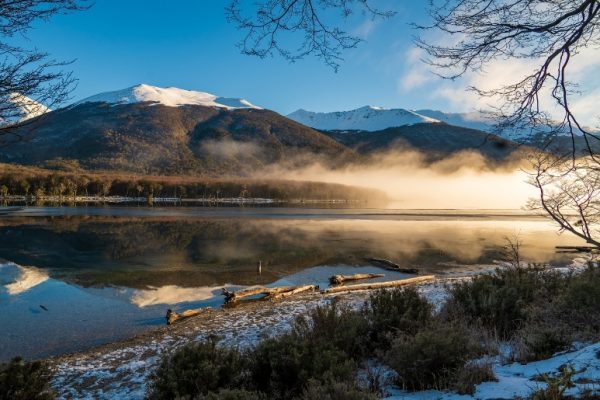 Una vista panorámica del Lago Escondido en Ushuaia al amanecer, mostrando las montañas andinas cubiertas de nieve profunda y bosques helados reflejados en el agua serena, con troncos flotando cerca de la orilla nevada y matorrales.