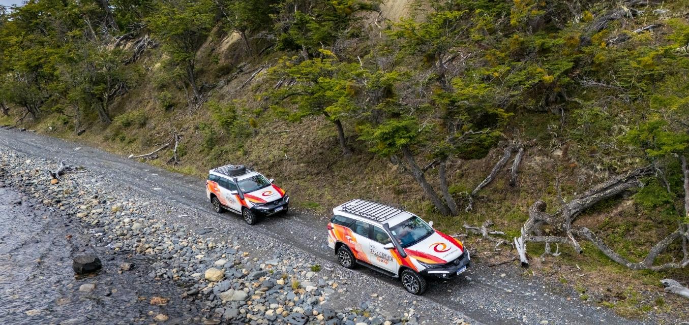 Dos camionetas 4x4 de Aswalek Expediciones circulando por la costa de piedras del Lago Fagnano, con el agua cristalina y el bosque de lengas bajo las montañas de Ushuaia al fondo.