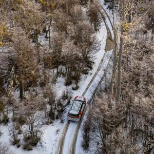 Una toma aérea cenital captura una camioneta blanca 4x4 conduciendo por un camino de tierra cubierto de nieve profunda, serpenteando a través de un denso bosque de lengas helado en un valle andino cerca de Ushuaia, bajo un cielo cubierto.