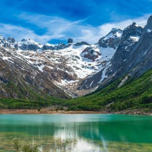 Paisaje panorámico de la Laguna Esmeralda en Ushuaia, mostrando sus aguas de color verde turquesa rodeadas de un bosque de lengas y las imponentes montañas nevadas de la Cordillera de los Andes bajo un cielo azul despejado.