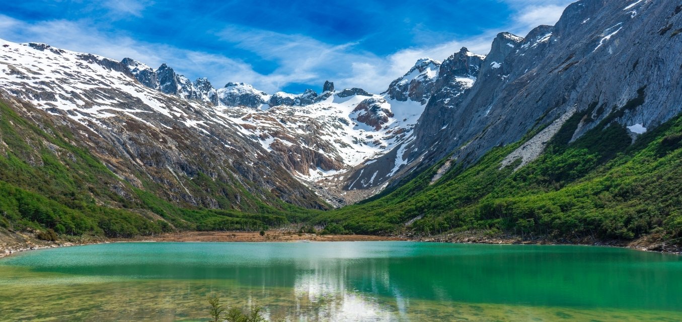 Paisaje panorámico de la Laguna Esmeralda en Ushuaia, mostrando sus aguas de color verde turquesa rodeadas de un bosque de lengas y las imponentes montañas nevadas de la Cordillera de los Andes bajo un cielo azul despejado.
