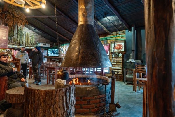 Vista interior del restaurante rústico de Las Cotorras en Ushuaia, mostrando el gran hogar central de ladrillo con chimenea de metal ardiendo, turistas relajándose, mesas de madera y decoración tradicional de montaña bajo un techo de madera inclinada.