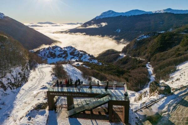 Una vista aérea captura a un grupo de turistas en la plataforma de vidrio y metal del Mirador Paso Garibaldi en invierno, Ushuaia, rodeados de montañas andinas cubiertas de nieve profunda y un valle lleno de niebla densa bajo el sol brillante.