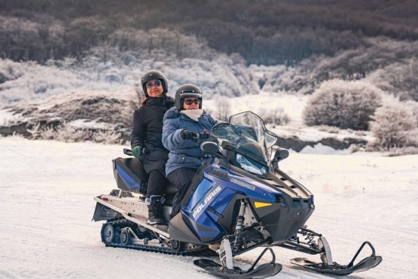 Dos turistas con cascos, anteojos de sol y ropa de invierno conducen una moto de nieve azul Polaris Advance en fila india por un camino cubierto de nieve profunda en un valle andino de Ushuaia, bajo un cielo nublado parcial con bosques de lengas helados al fondo.