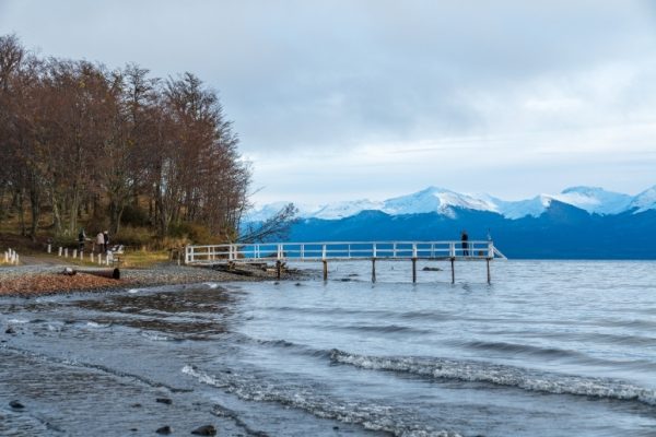 Un muelle de madera blanco con una barandilla se extiende hacia el Lago Fagnano en Ushuaia en un día nublado, con montañas andinas nevadas al fondo y matorrales invernales en la orilla de guijarros.