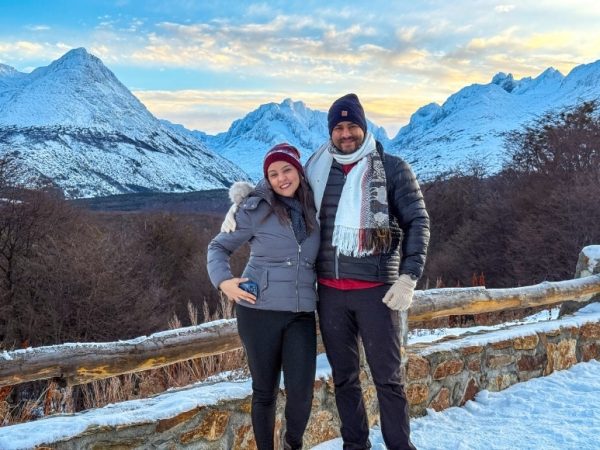 Una pareja de turistas vestida con ropa de invierno técnica posa sonriente frente a la cordillera de los Andes nevada y el Valle Carbajal en Ushuaia, Argentina, en un mirador con una barandilla de madera rústica y nieve profunda.