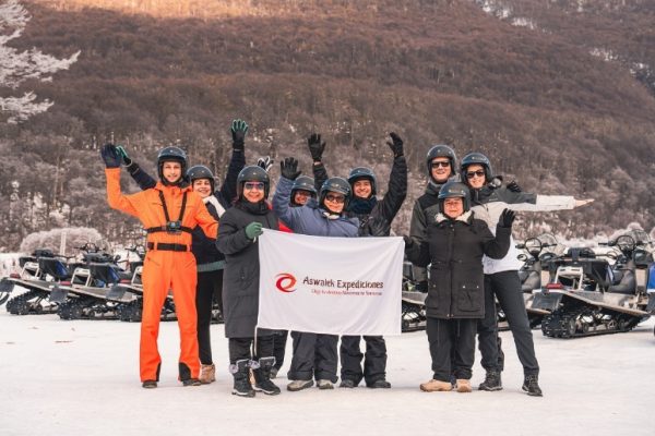 Un grupo de turistas con cascos y ropa de nieve de invierno posa sonriendo y saludando en un valle andino de Ushuaia. Dos de ellos sostienen una bandera blanca con el logo de Aswalek Expediciones. Motos de nieve azules y blancas están estacionadas a la izquierda y derecha. Al fondo, un bosque de lengas helado y montañas nevadas bajo un cielo nublado parcial.