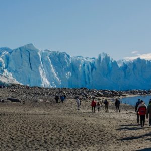 Un grupo de turistas camina por una playa de arena oscura hacia un enorme frente de glaciar azul bajo un cielo despejado.