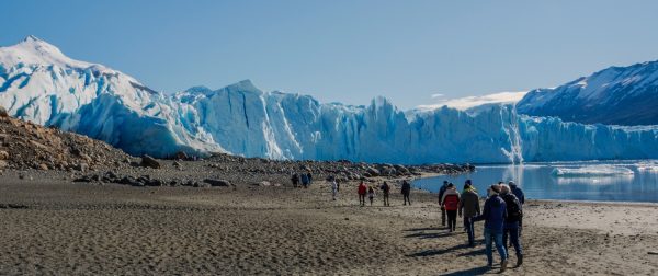 Un grupo de turistas camina por una playa de arena oscura hacia un enorme frente de glaciar azul bajo un cielo despejado.