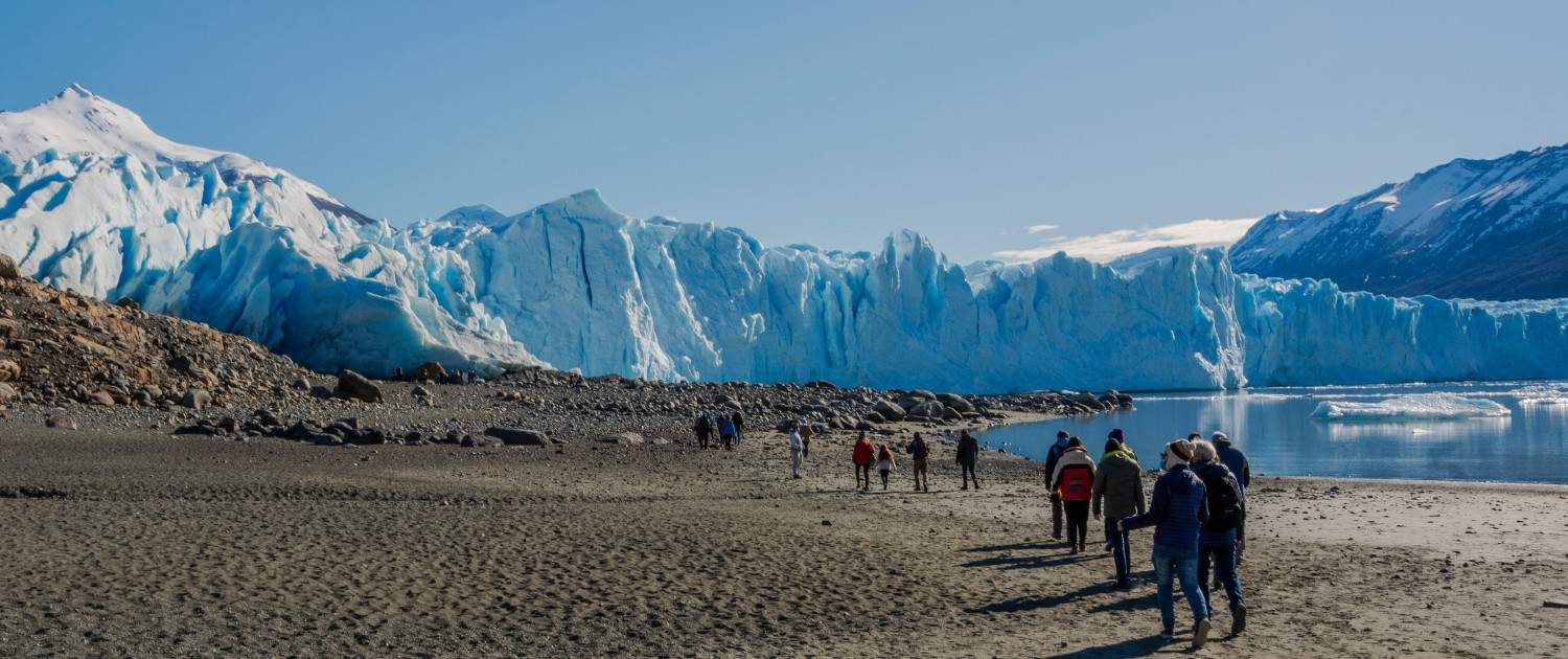 Un grupo de turistas camina por una playa de arena oscura hacia un enorme frente de glaciar azul bajo un cielo despejado.