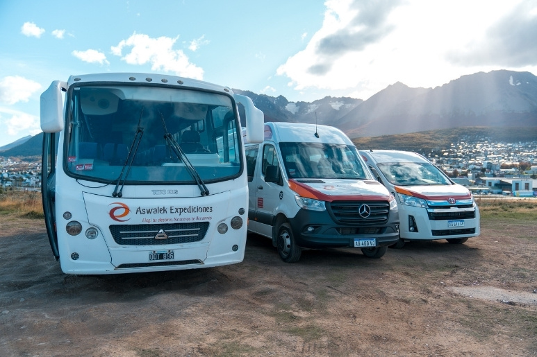 Una toma frontal de nivel medio que captura la flota de vehículos de "Aswalek Expediciones", incluyendo un ómnibus blanco grande, una van Mercedes-Benz Sprinter blanca y otra furgoneta Toyota blanca, estacionadas en un camino de tierra frente a las montañas nevadas de la Patagonia bajo un cielo soleado y nublado.