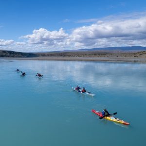 Grupo de turistas en kayaks dobles y simples navegando en fila por las aguas tranquilas y turquesas del Lago Argentino bajo un cielo parcialmente nublado.