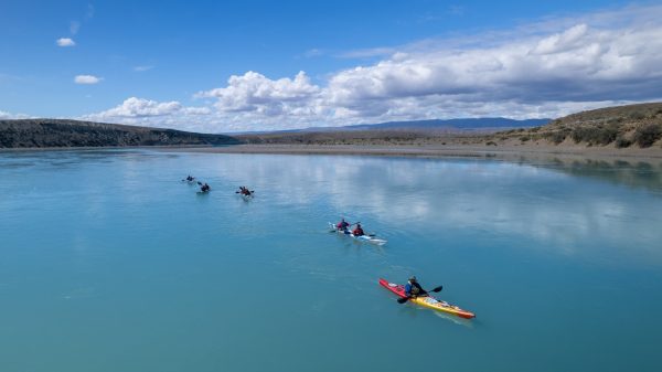 Grupo de turistas en kayaks dobles y simples navegando en fila por las aguas tranquilas y turquesas del Lago Argentino bajo un cielo parcialmente nublado.