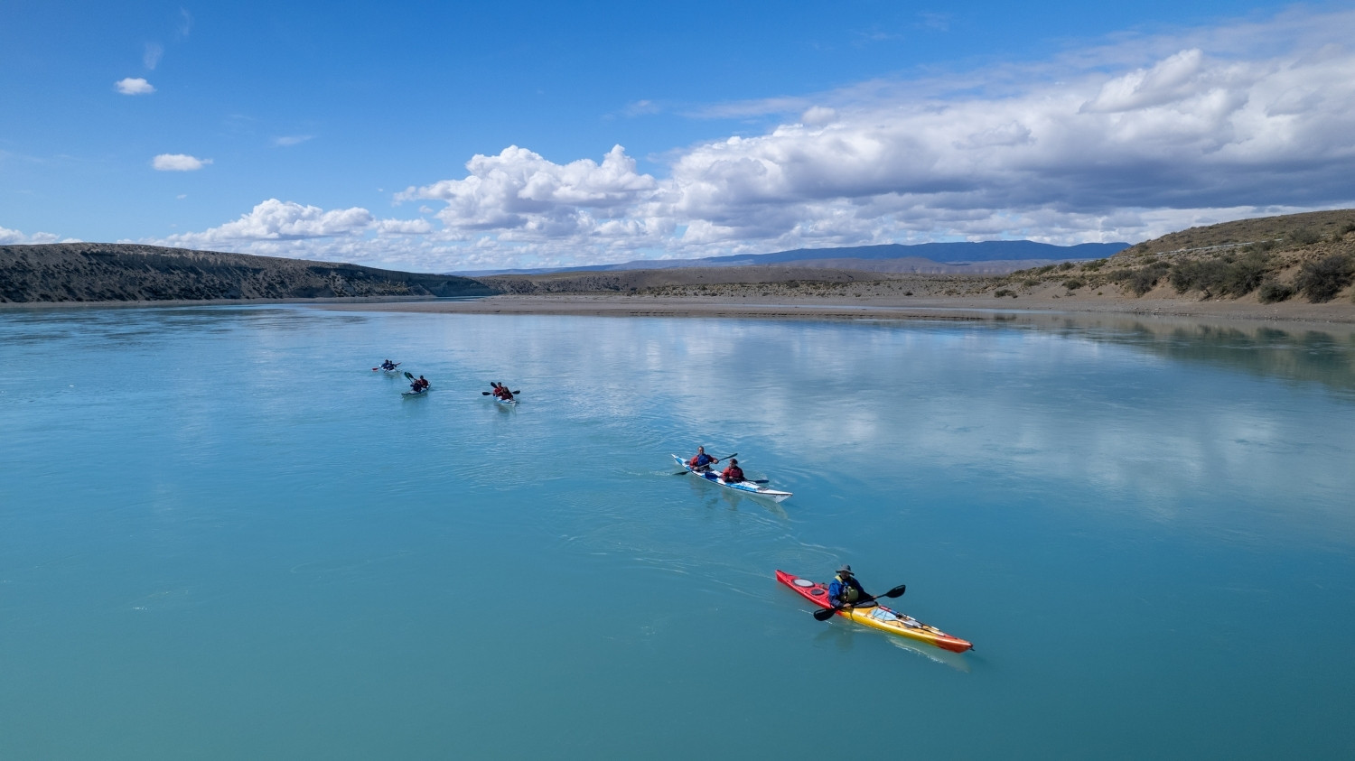 Grupo de turistas en kayaks dobles y simples navegando en fila por las aguas tranquilas y turquesas del Lago Argentino bajo un cielo parcialmente nublado.