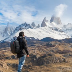 Un turista de espalda con mochila negra contemplando el imponente Cerro Fitz Roy nevado desde una cumbre rocosa sobre el valle.