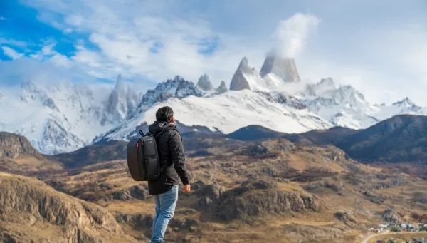 Un turista de espalda con mochila negra contemplando el imponente Cerro Fitz Roy nevado desde una cumbre rocosa sobre el valle.