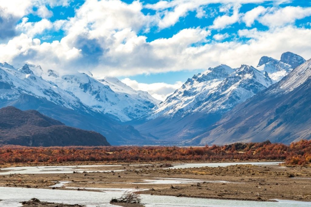 Paisaje panorámico de un río serpenteante en un valle rodeado de bosques otoñales y cordilleras con picos nevados bajo un cielo nublado.