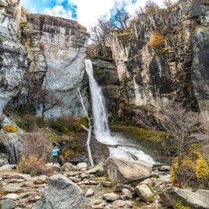 Una gran cascada de agua cristalina cayendo entre imponentes paredes de roca, con una persona fotografiando el paisaje en la base.