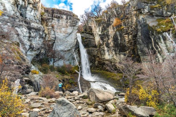 Una gran cascada de agua cristalina cayendo entre imponentes paredes de roca, con una persona fotografiando el paisaje en la base.