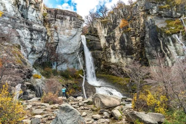 Una gran cascada de agua cristalina cayendo entre imponentes paredes de roca, con una persona fotografiando el paisaje en la base.